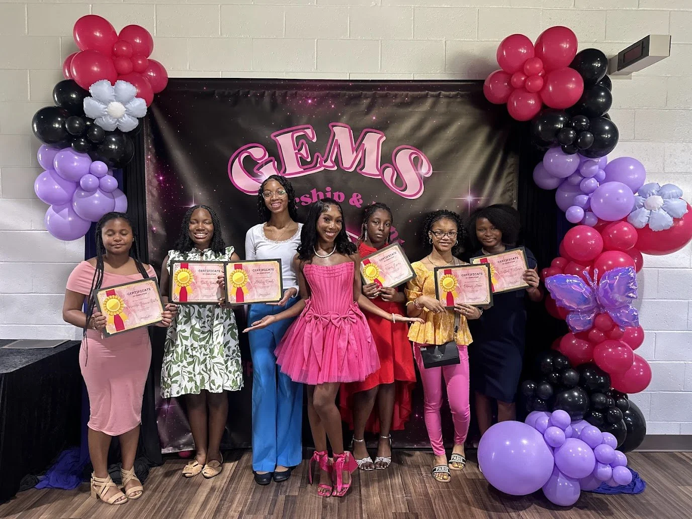Group of seven young girls and women standing in front of a backdrop that reads 'GEMS' with balloons and decorations. They are holding certificates of achievement and dressed in colorful outfits for an award ceremony or celebration.