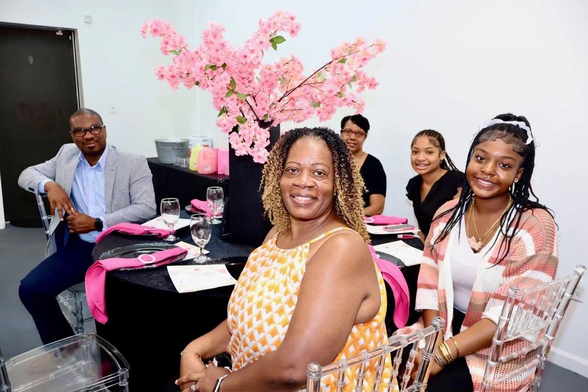 Group of five people sitting at a decorated table with pink napkins, glasses, and a large pink flower arrangement, smiling indoors.