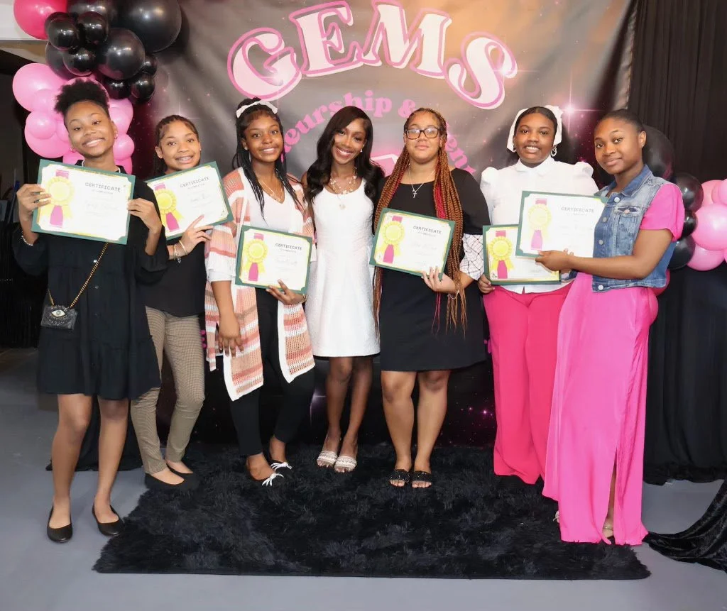 Group of seven women and girls standing together, holding certificates at an event with a backdrop reading 'GEMS Leadership & Scholarship'.