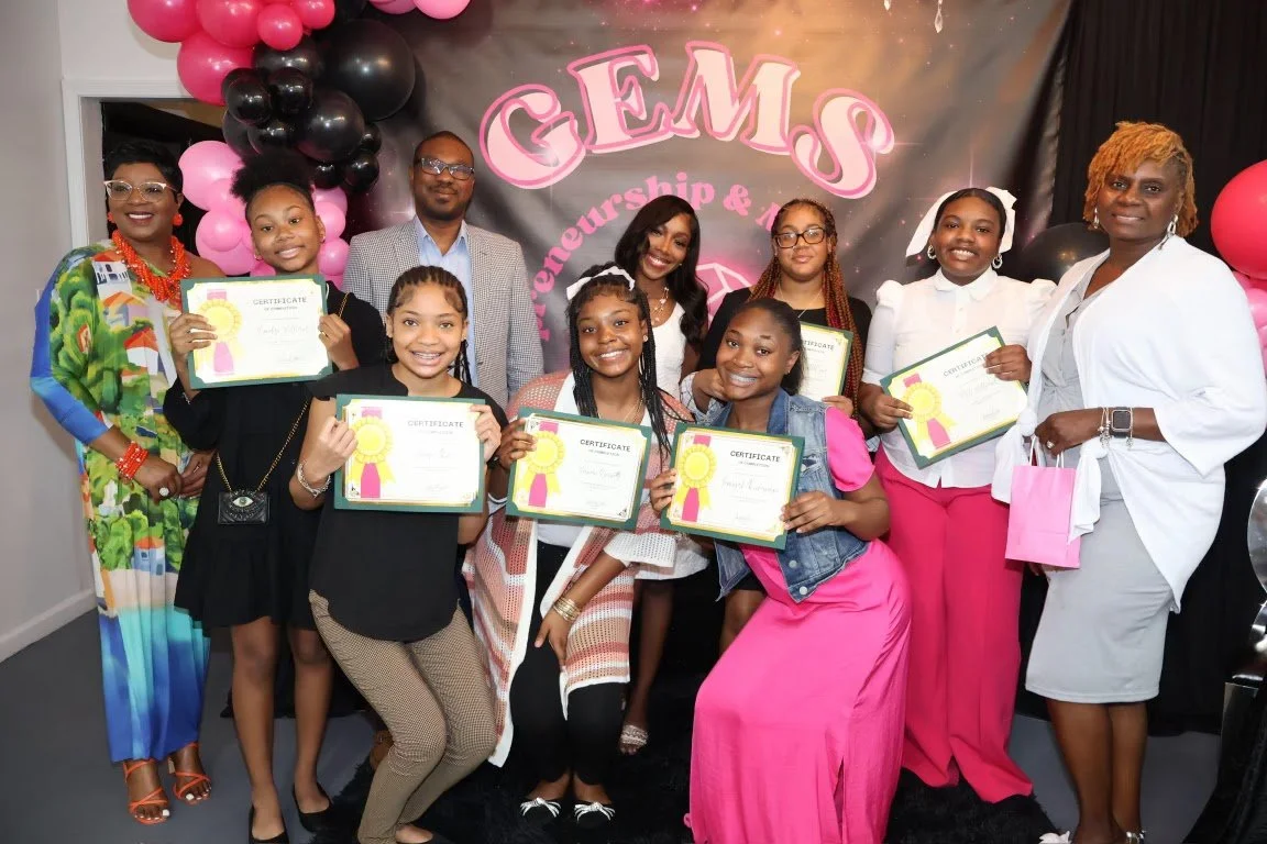 Group of girls and women holding certificates at an event with a 'GEMS' banner and pink, black, and gold balloons.