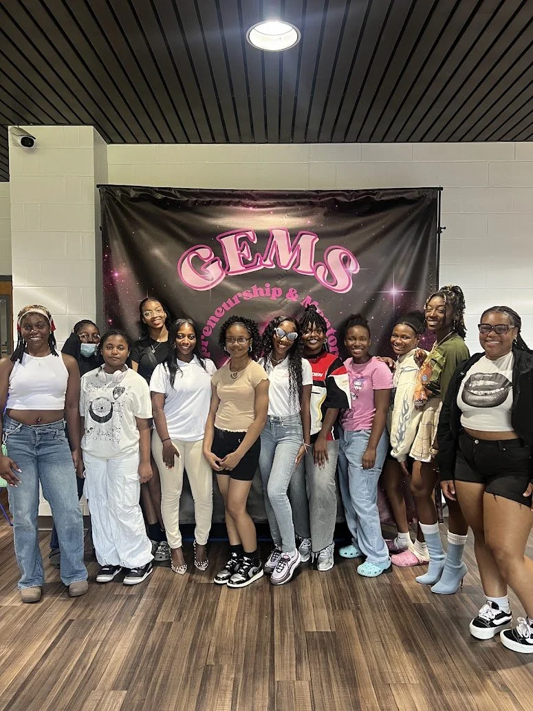Group of young women standing in front of a GEMS banner at an entrepreneurship and mentorship event, smiling and posing for the photo.