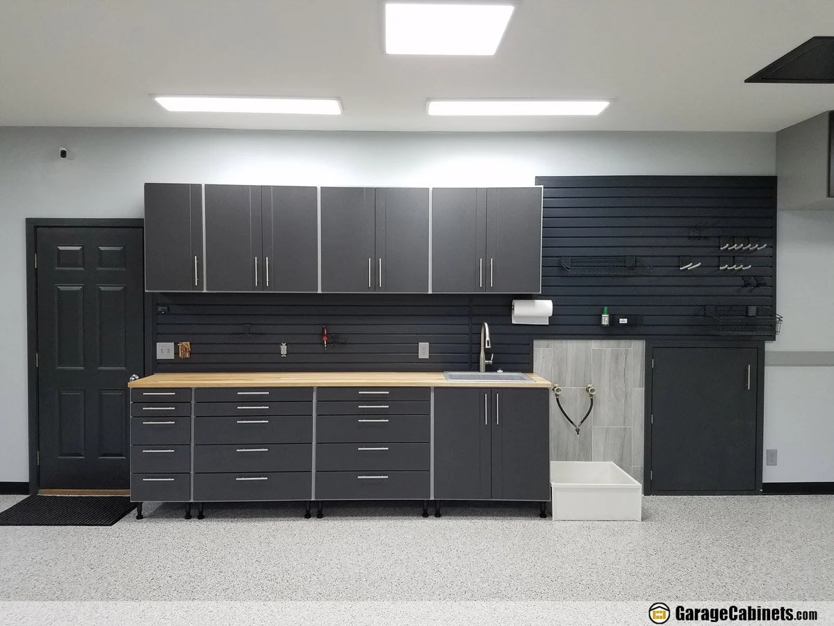 Garage with black cabinets, gray upper cabinets, black slat wall with organized tools, gray tiled mudroom area, white utility sink, ceiling lights, and neutral speckled flooring.