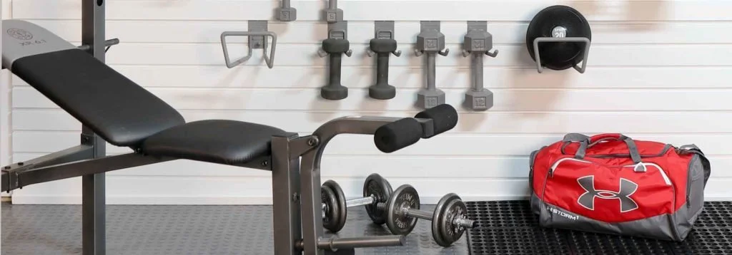 A gym workout area with a black adjustable workout bench, a red Under Armour sports bag, a pair of dumbbells on the floor, and wall-mounted weights and equipment.