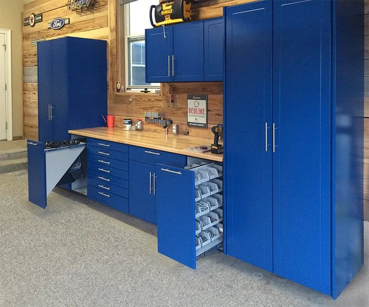Blue storage cabinets and drawers with a wooden workbench on a wall in a garage or workshop, containing tools, supplies, and storage bins.