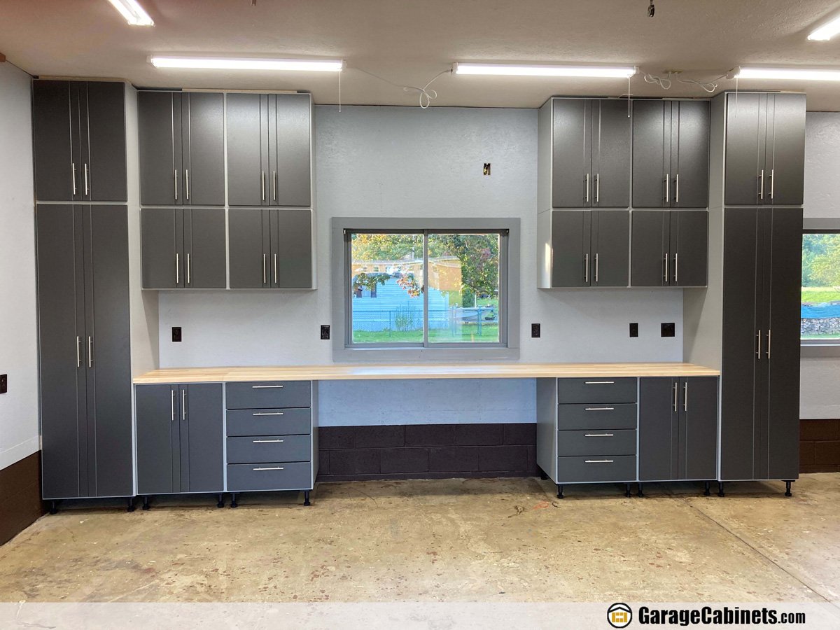 Modern gray garage cabinets with black hardware, a long white workbench, and a window showing a yard outside.