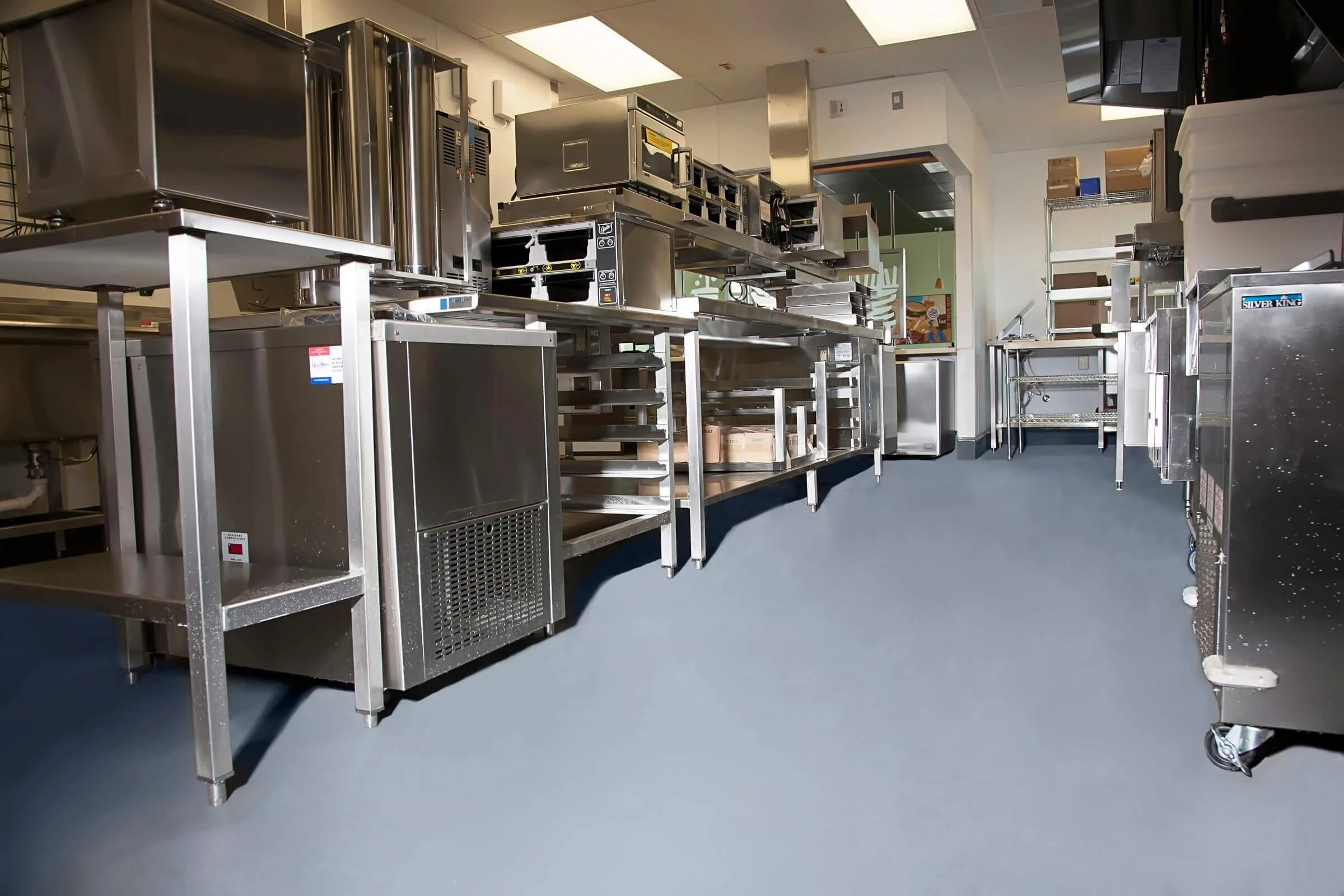 Industrial kitchen or restaurant supply room with stainless steel tables, appliances, and shelving units, some with boxes and kitchen equipment.