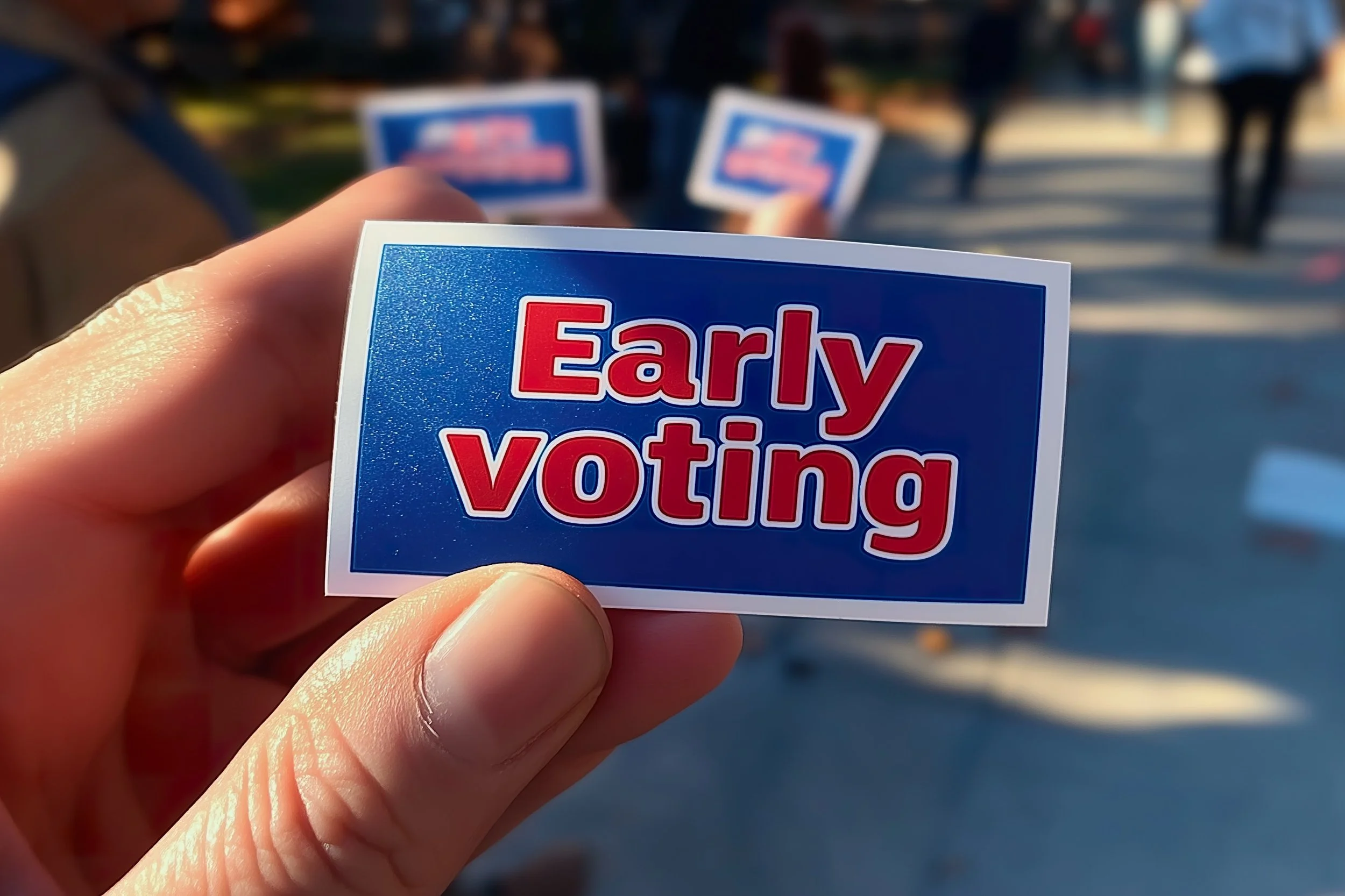 Close-up of a hand holding a small political sticker that reads 'Early Voting' in red and white text on a blue background, with additional similar stickers in the blurred background.