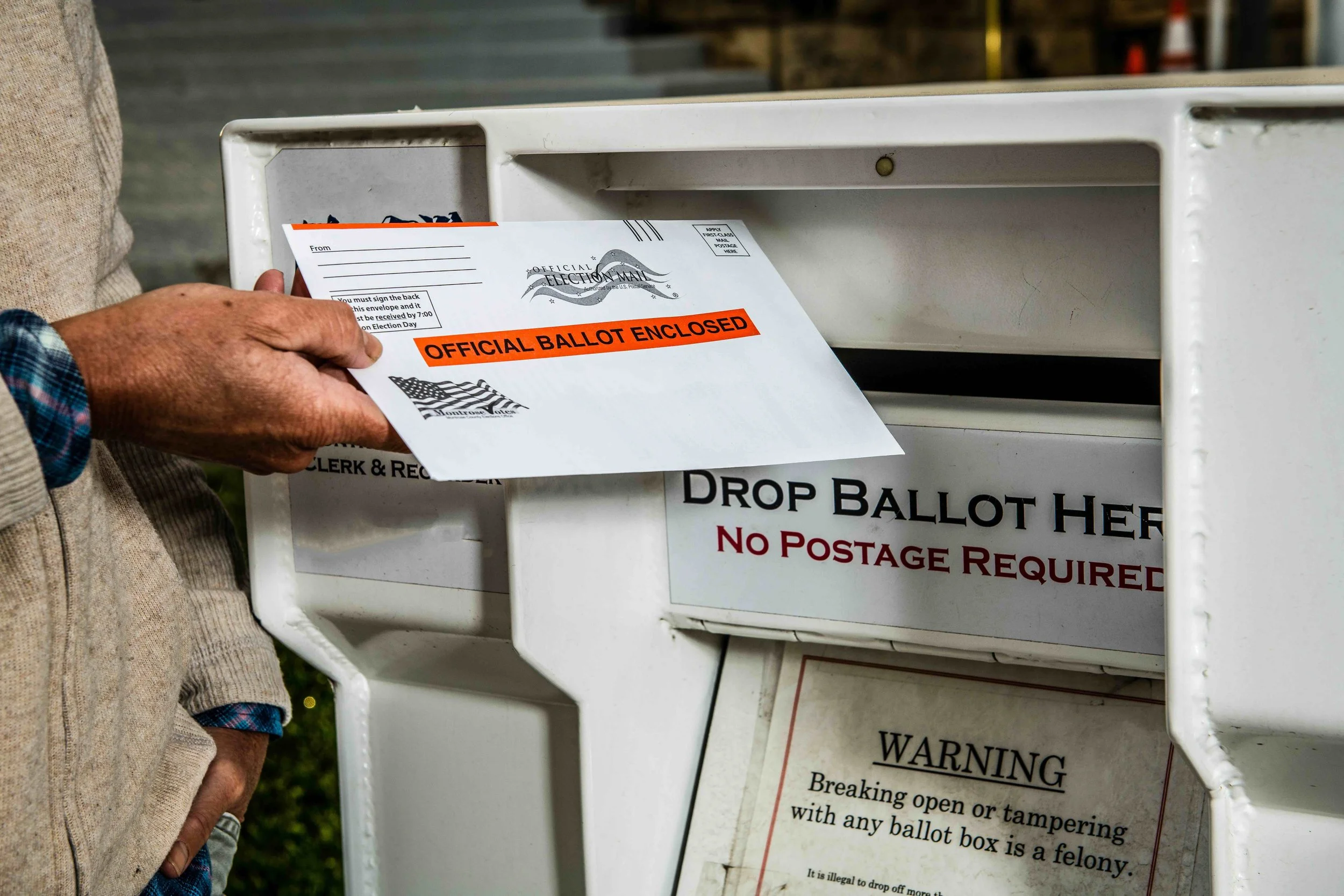 Close-up of a person dropping off an official ballot into a white drop box, with a sign indicating it is for official election mail and sealed with orange tape.