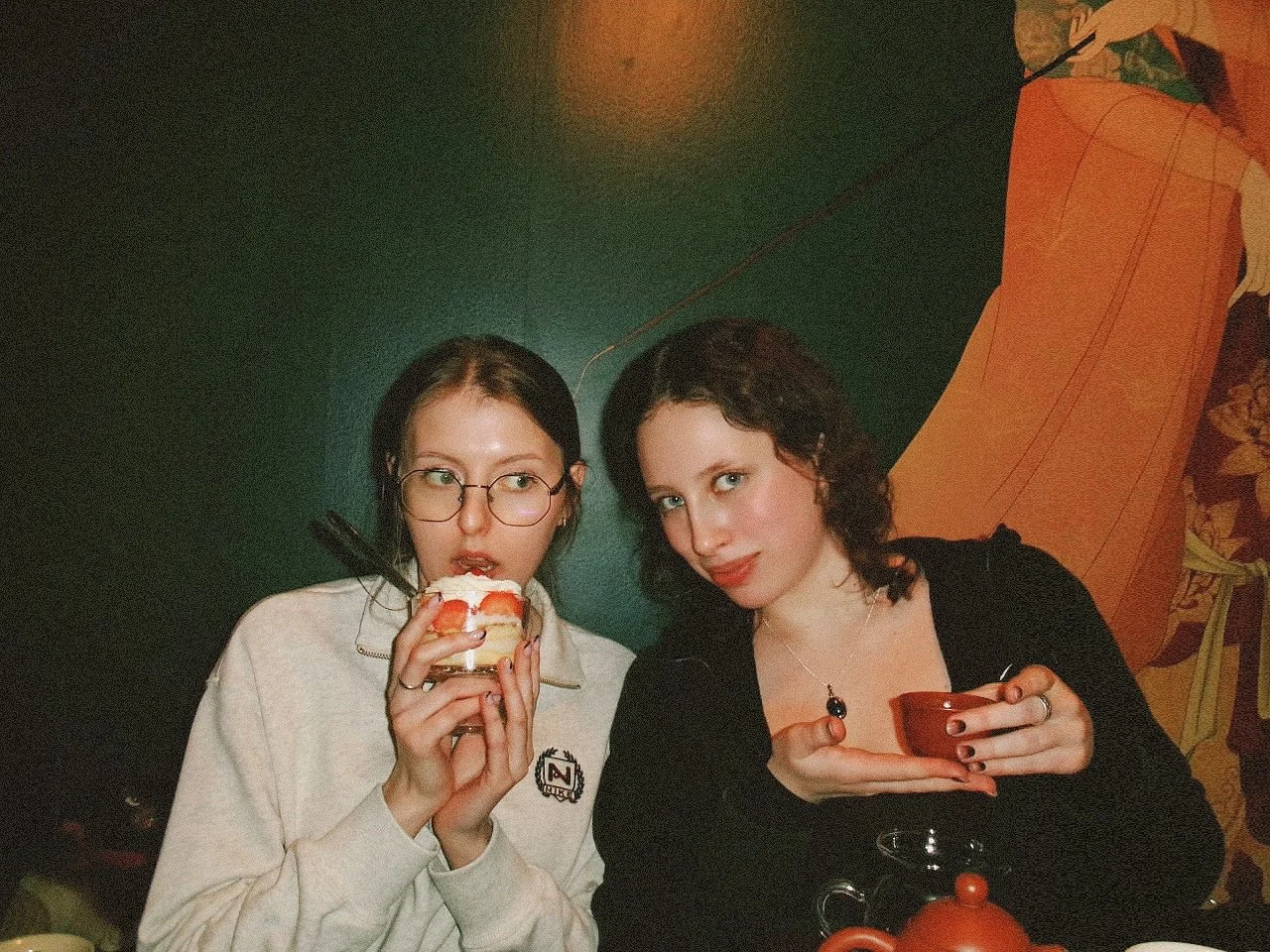 Two young women sitting at a table, one holding a slice of cake and the other holding a teacup, in a cozy indoor setting with a colorful mural or textured wall in the background. - Anna and Stanislava - founders of Symphony of views