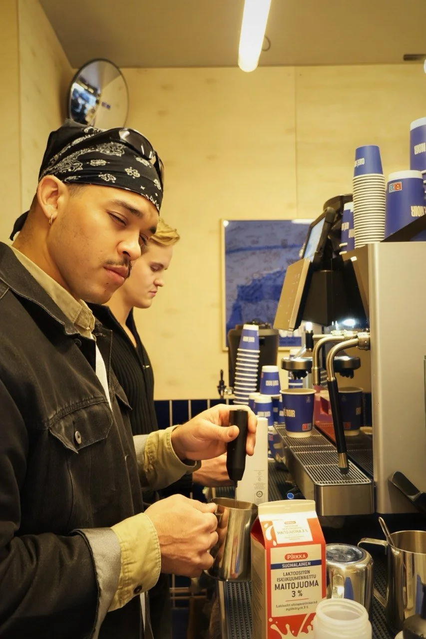 Two people preparing coffee at a cafe counter, with a barista holding a milk frothing pitcher and a steam wand, surrounded by cups, milk cartons, and coffee equipment.