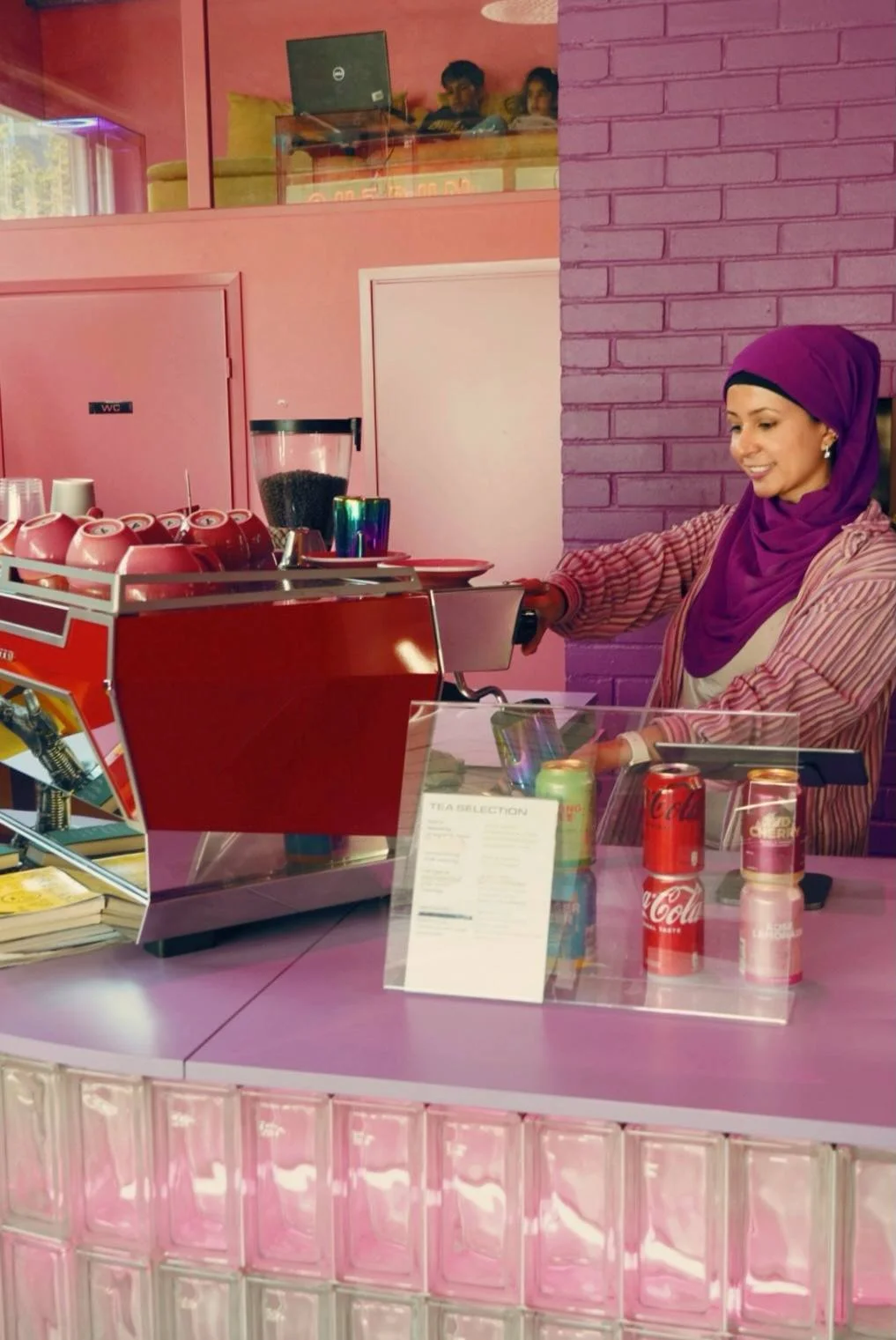 A woman wearing a purple headscarf and striped shirt ordering at a pink coffee shop counter with a pink brick wall, soda cans, and tea selection menu. - OUTRUN
