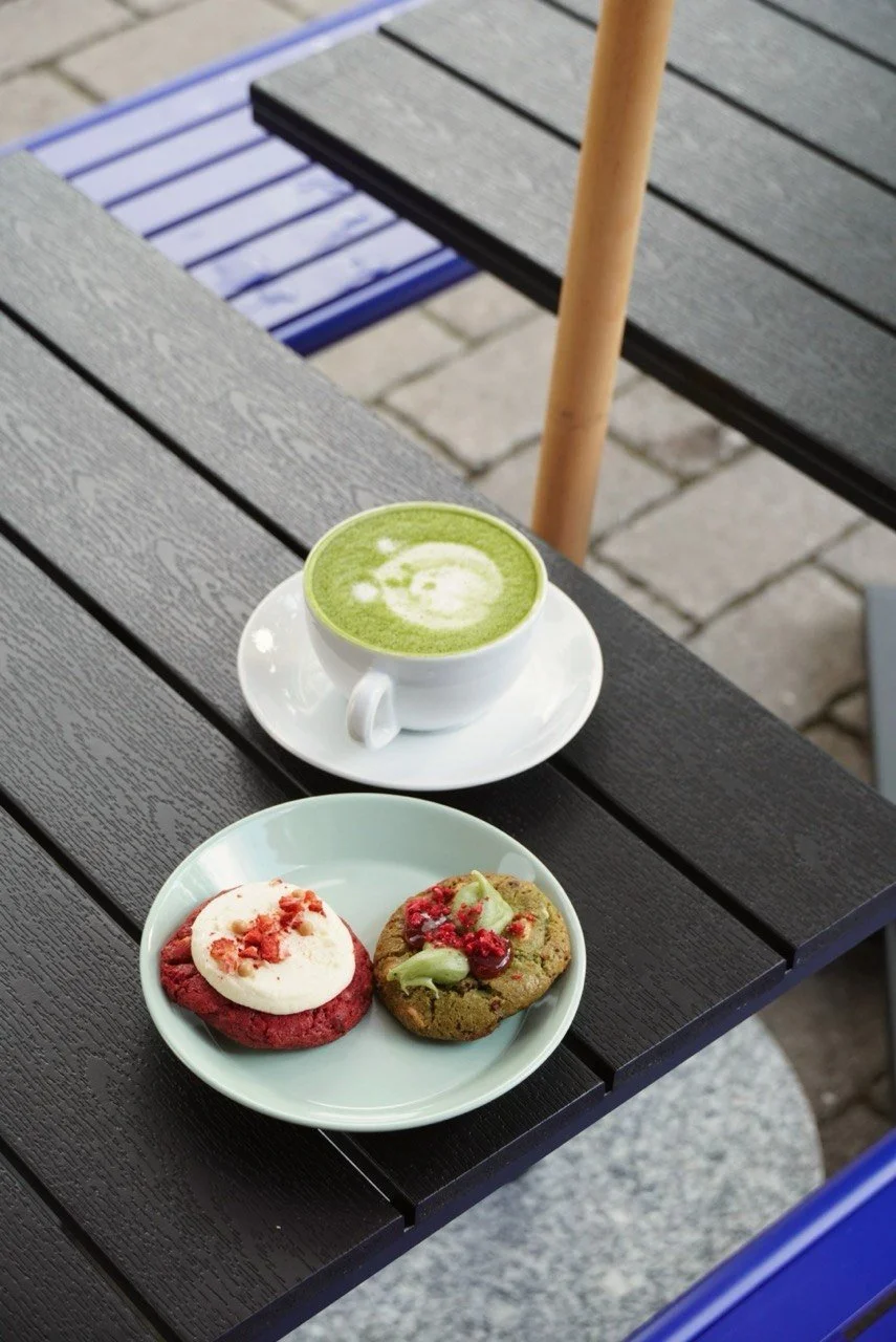 A white cup of green matcha latte with foam art, placed on a saucer on a black wooden outdoor table. In front of it, a light green plate holds two cookies—one red velvet with cream cheese frosting and crumb topping, and one chocolate chip with white drizzle and green filling.