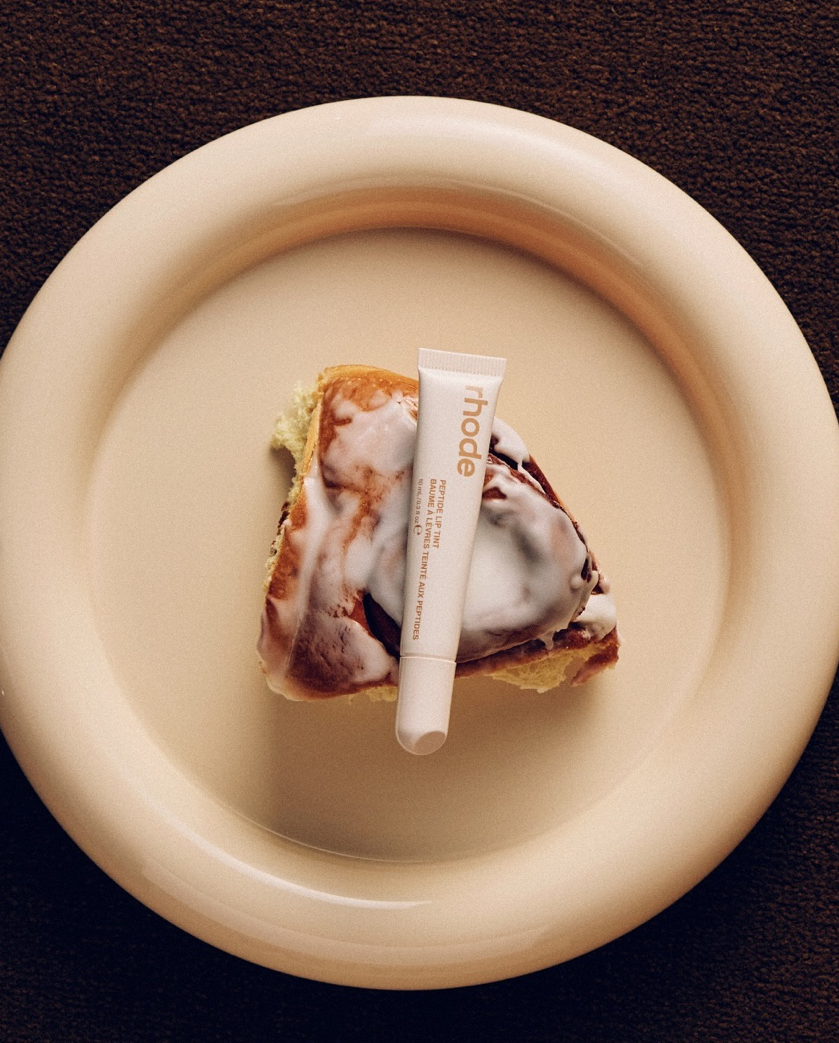 A slice of cake with icing and a tube of skincare product labeled 'rhode' placed on top, on a white plate.
