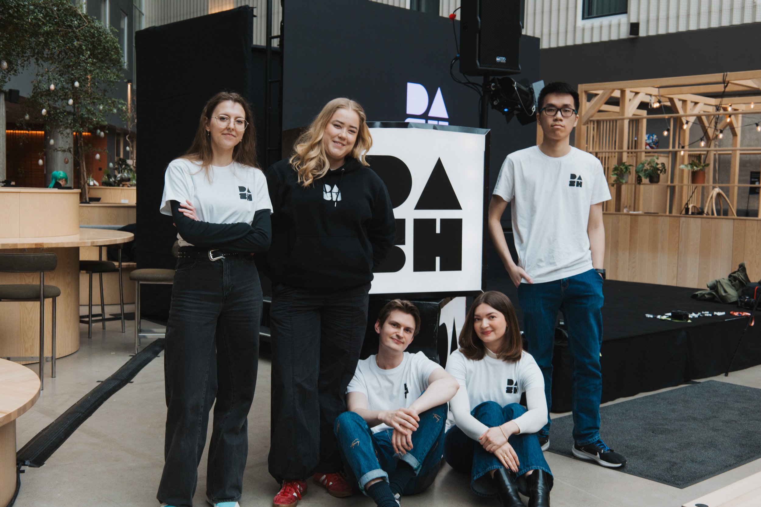Group of six young people, three standing and three sitting, posing for a photo indoors at a casual event or gathering, with a background featuring a large screen, speakers, and wooden decor.