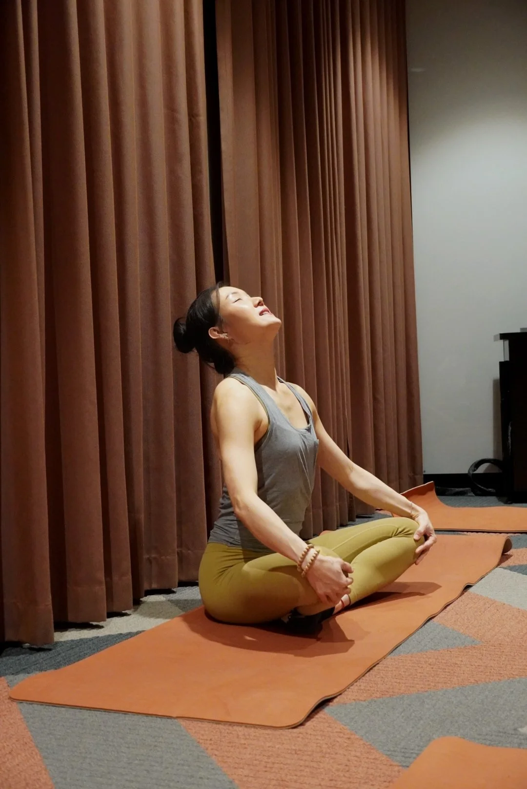 A woman practicing yoga or meditating on a mat in a room with brown curtains, sitting cross-legged with her hands resting on her knees and eyes closed.