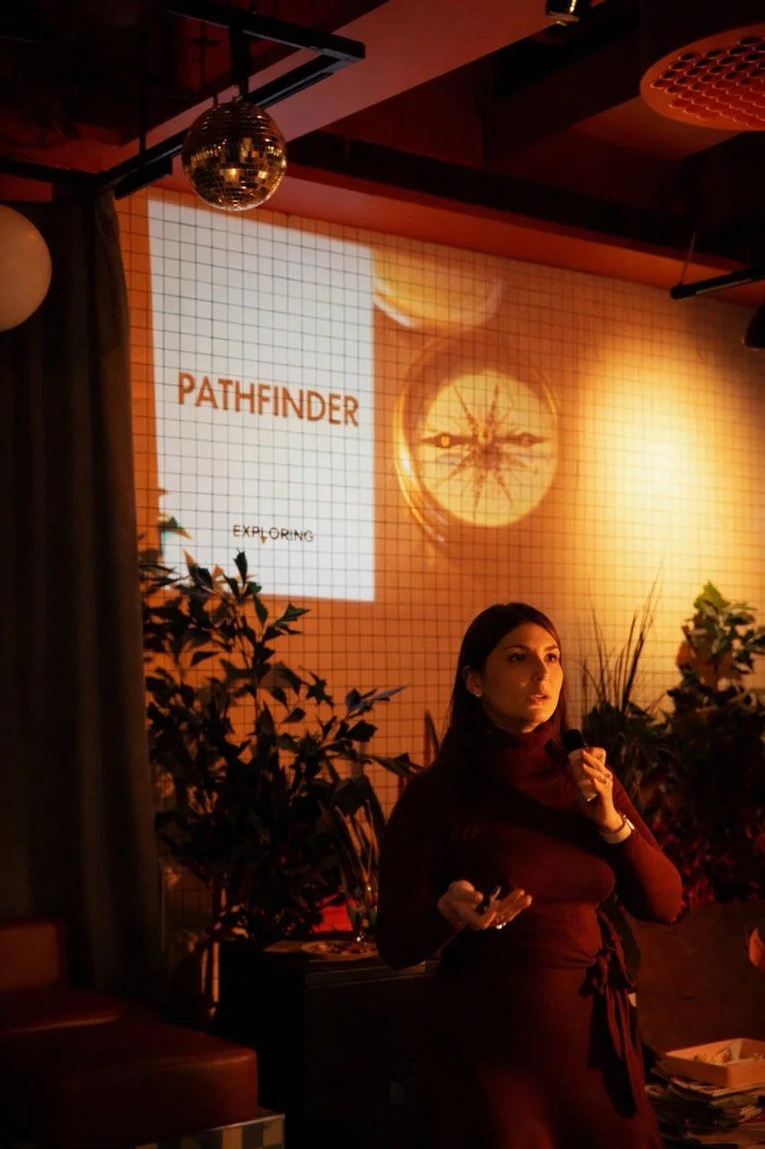 A woman giving a presentation in a dimly lit room with plants, a screen displaying the words 'PATHFINDER' and 'EXPLORING,' and a disco ball hanging from the ceiling.