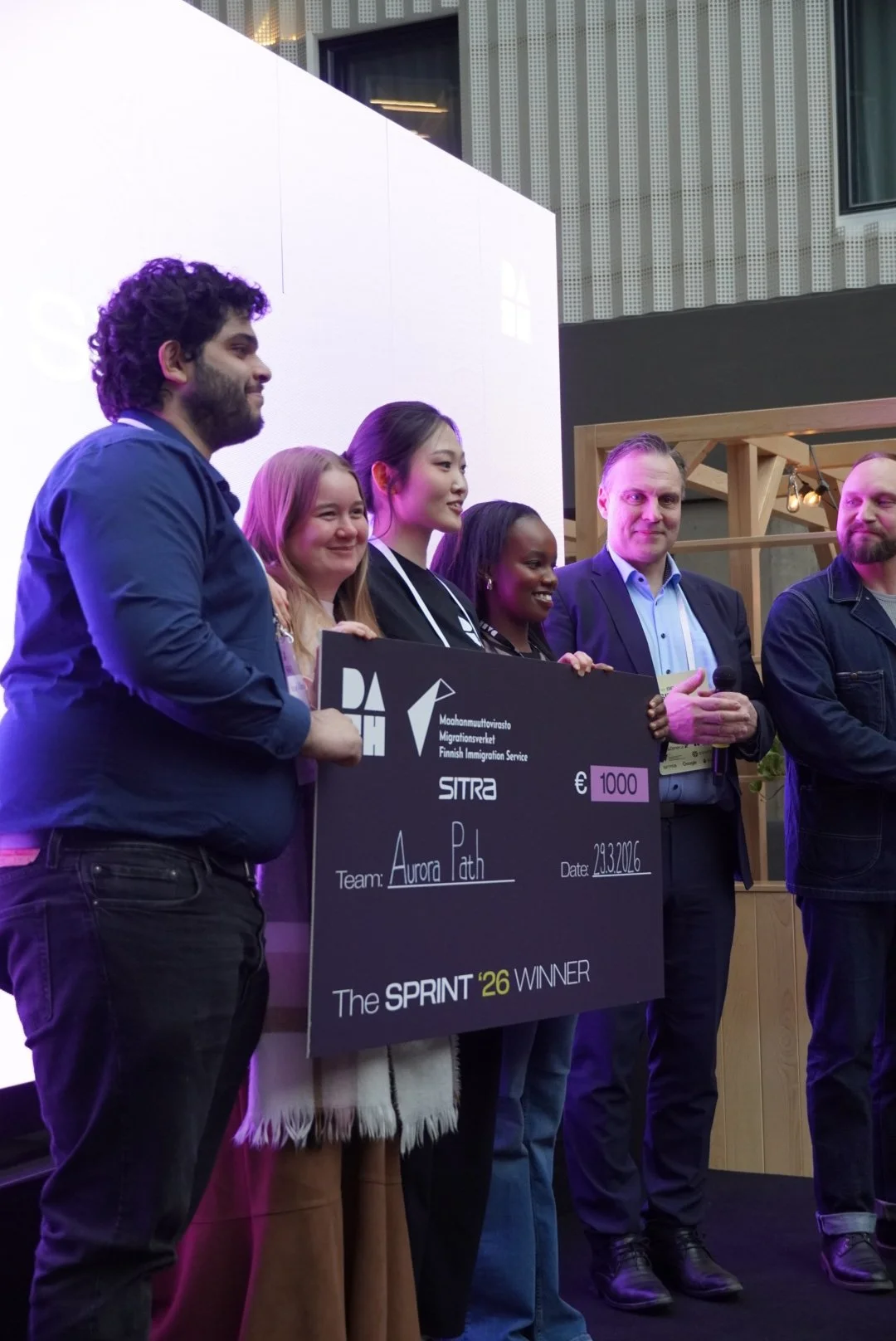Group of five people holding a large check on stage at an event, celebrating a win, with people smiling and standing in front of a white large screen and a wooden decorative structure.