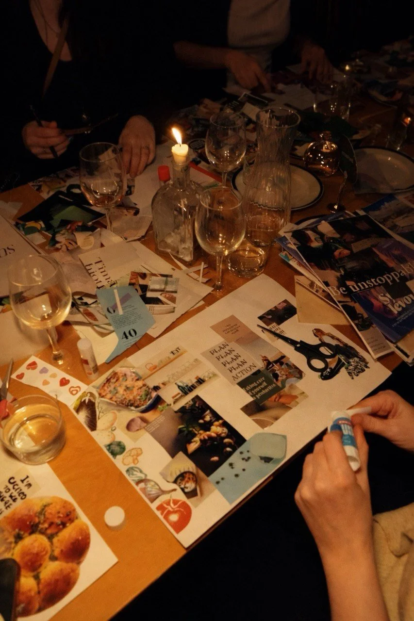 People around a table engaged in a creative activity, organizing and working with magazines, scissors, and glue, with several glasses of wine and a lit candle in a dimly lit setting.
