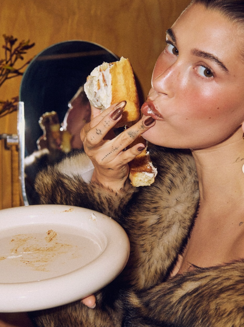 A young woman with tattoos and a fur coat is holding a plate with leftover food while eating a slice of cake with her fingers, and looking into the camera. A mirror reflects her profile.