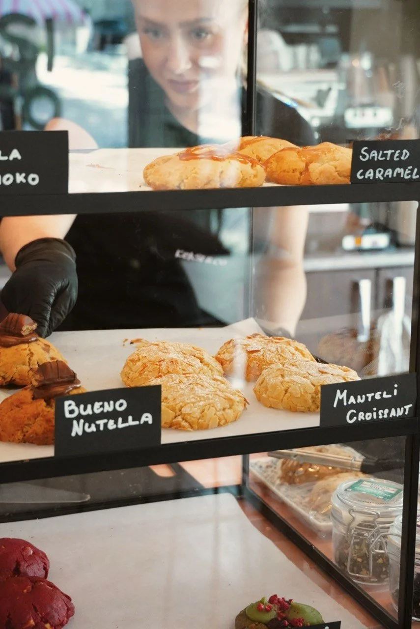 An assortment of cookies and pastries displayed behind a glass case, with a woman in the background looking at them. The visible labels include "Buono Nutella," "Manteli Croissant," and salted caramel.