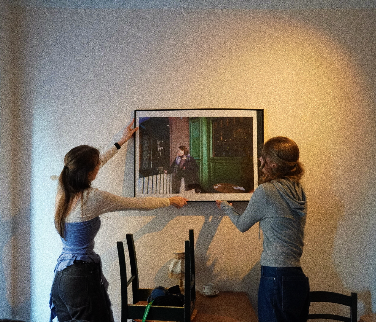 Two women hanging a framed artwork on a wall in a room with a table and chairs.