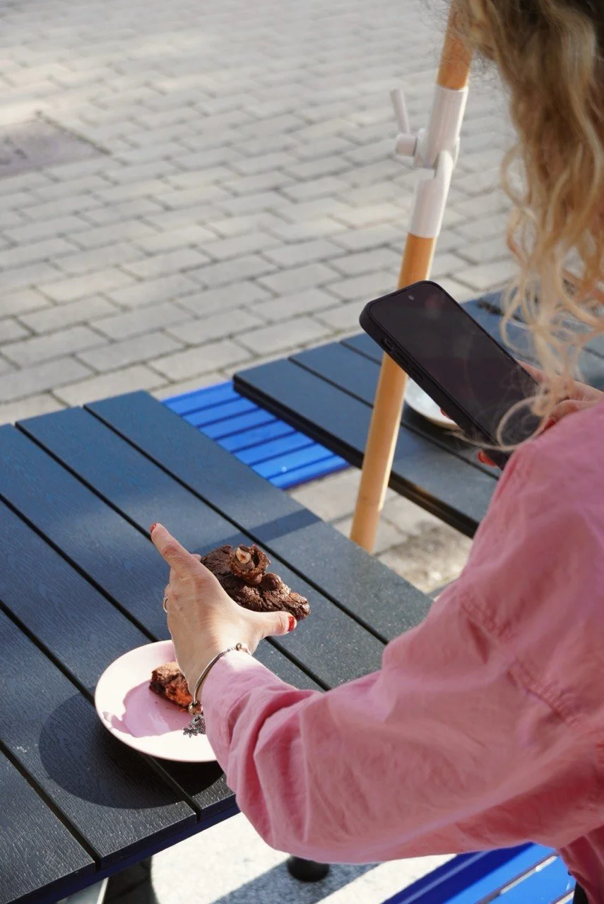 A woman with curly blonde hair in a pink shirt is taking a photo of a chocolate brownie with her smartphone at an outdoor cafe table.