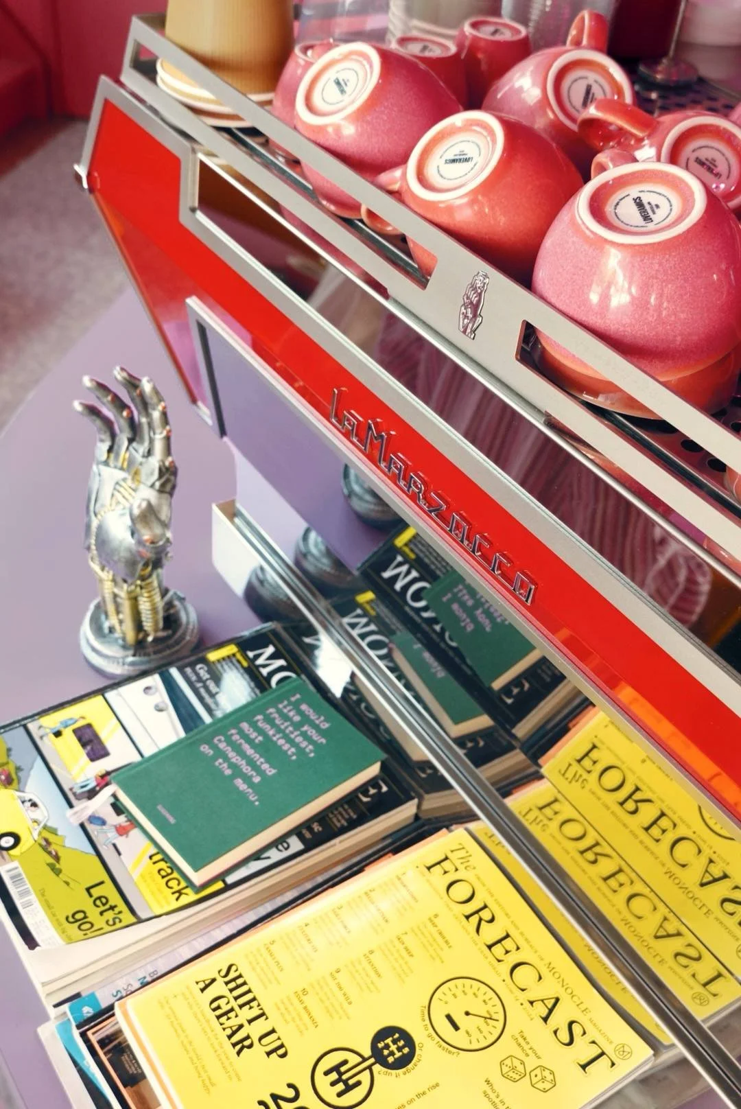 A display in a bookstore with red mugs, a sculpture of a robotic hand, and several books including 'The Forensic' and others titled in Greek.