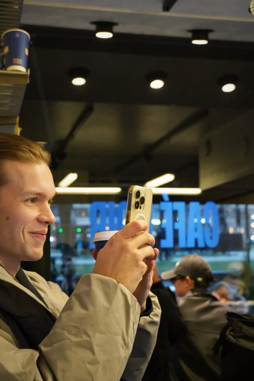 A young man with short hair smiling while holding a smartphone and a cup in a cafe.