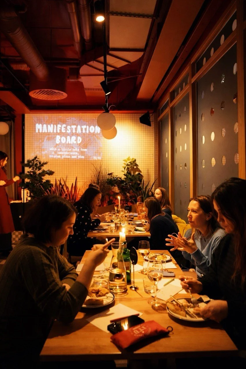 People sitting at a long dining table during a gathering, with a presentation screen in the background displaying 'Manifestation Board' and candles on the table.