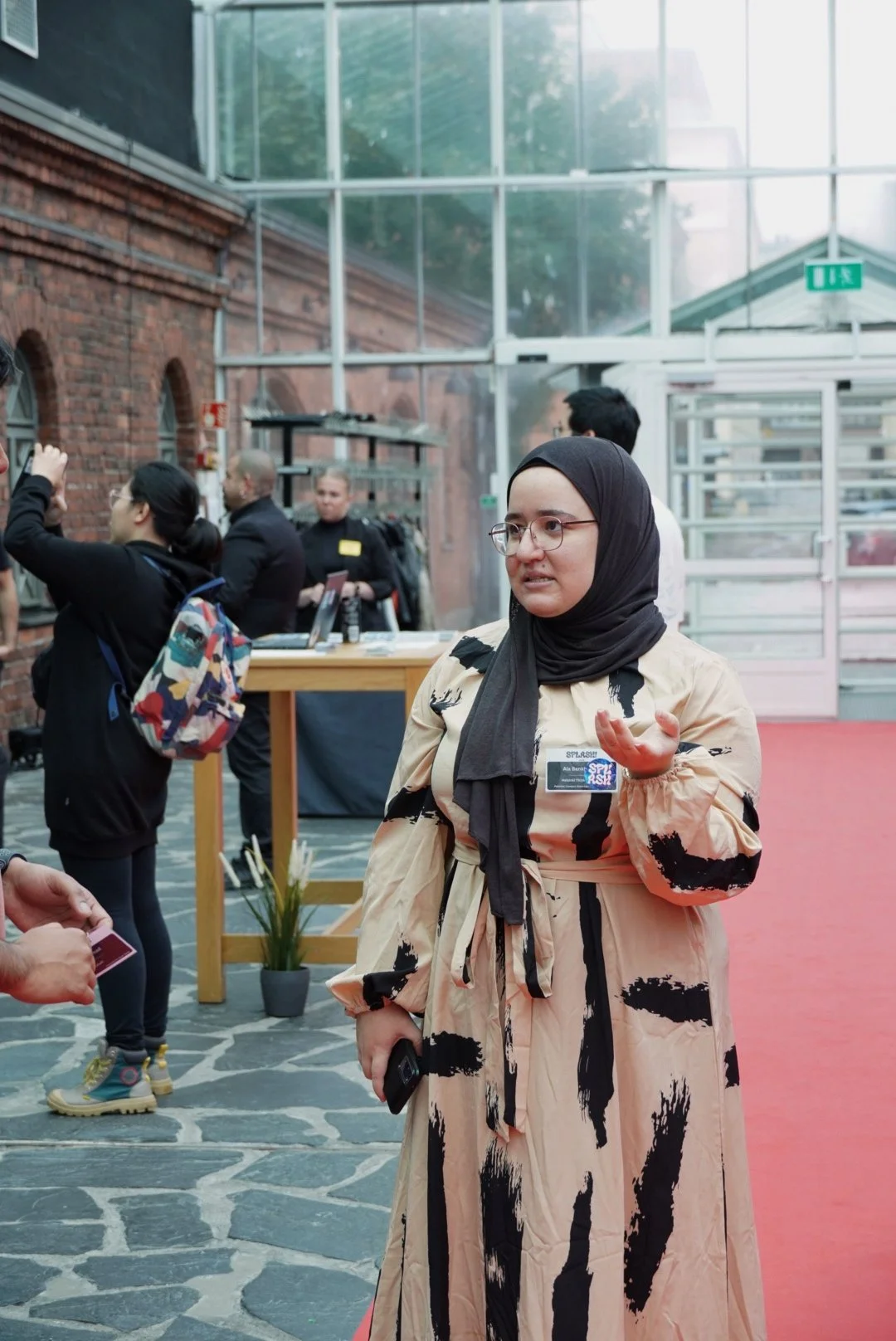 A woman wearing a beige dress with black abstract patterns and a black hijab, holding a phone, standing on a red carpet in an indoor venue with a glass ceiling and brick walls. Several people are in the background, some taking photos and some at a reception desk.
