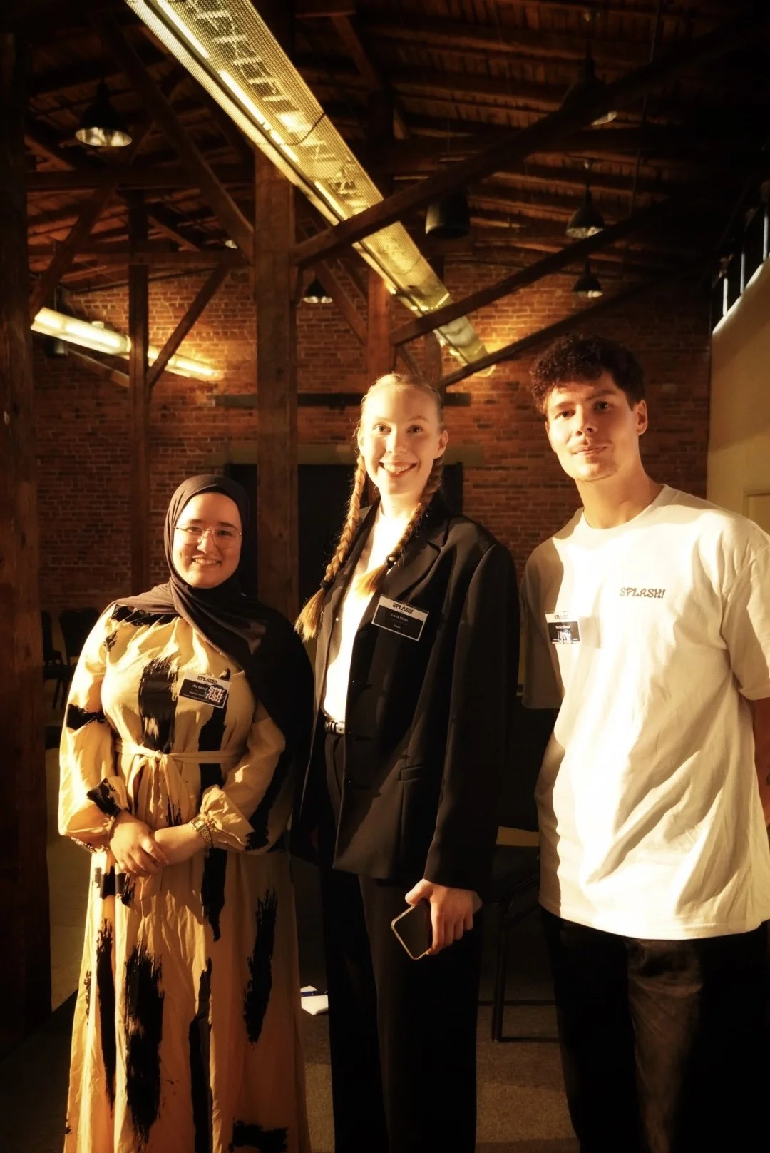 Three young people, two women and one man, standing together in a warmly lit indoor setting with exposed brick walls and wooden beams. They are smiling and wearing conference name tags.