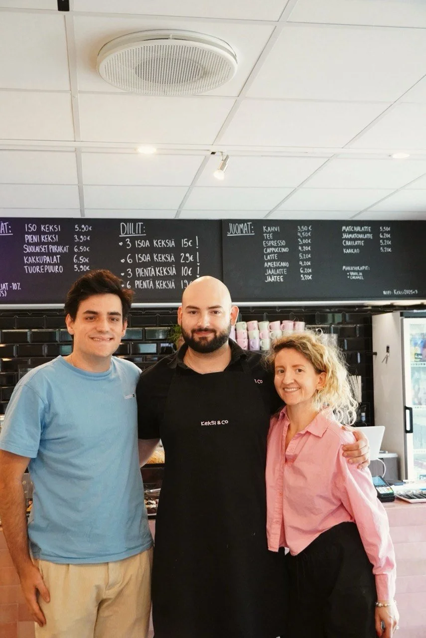 Three smiling people, two men and a woman, standing inside a cafe behind the counter, with a blackboard menu above them.