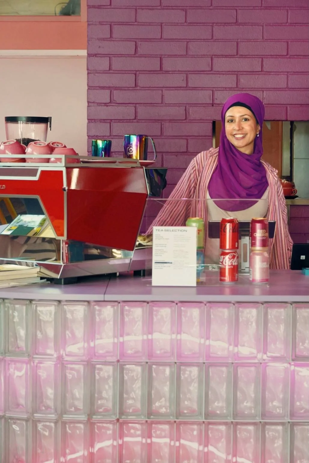 A woman standing behind a pink counter with a pink tiled front, smiling at the camera. She is wearing a purple headscarf and striped pink jacket. There are soda cans and a menu on the counter, with a pink brick wall in the background.