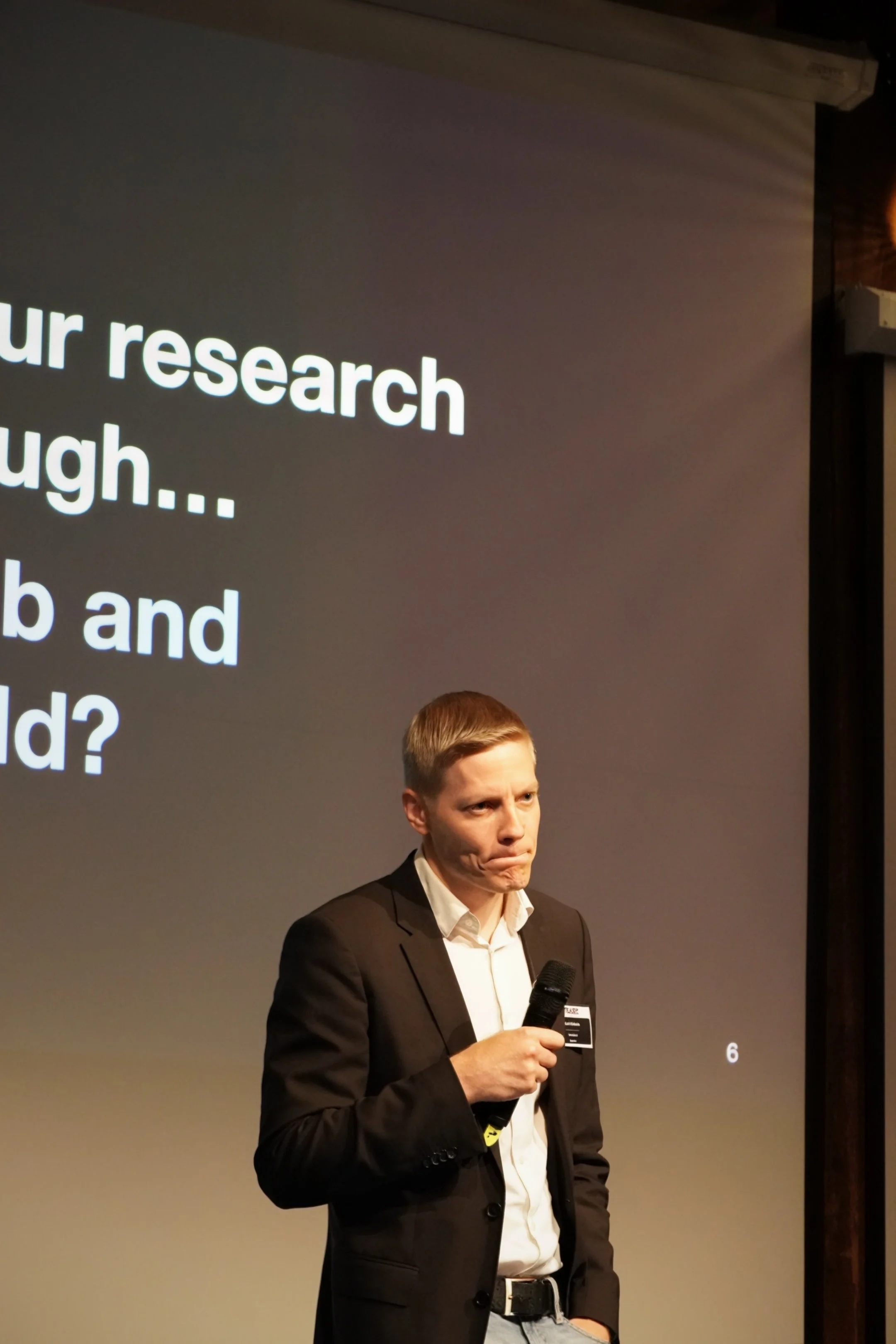 A man in a black suit and white shirt, holding a microphone, standing in front of a presentation screen with partially visible text.