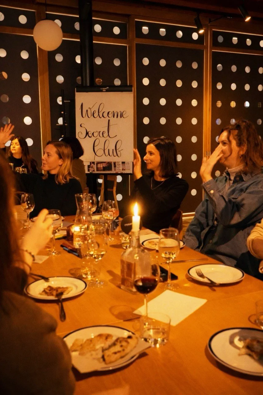 Group of women gathered around a dining table during a social event, with a sign that says 'Welcome Secret Club' in the background, in a warmly lit room with polka dot wall decor.