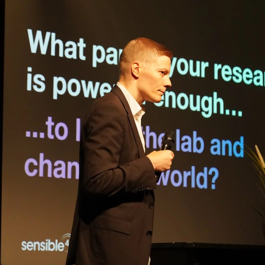 A person in a black suit holding a microphone, standing in front of a presentation slide with the text, 'What part of your research is powerful enough... to change the world?'