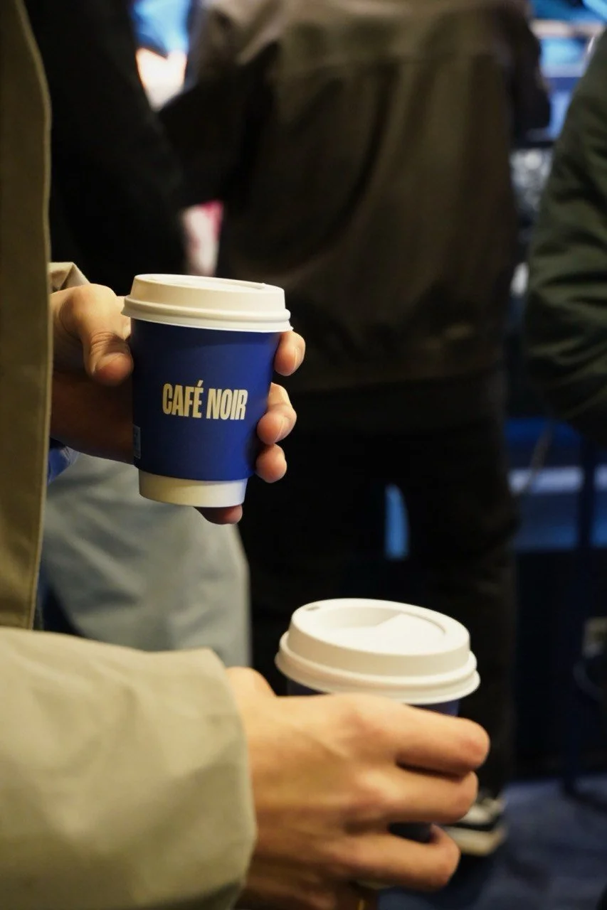 Close-up of two people holding takeaway coffee cups, one with a blue sleeve labeled 'CAFÉ NOIR,' in a busy café or coffee shop.