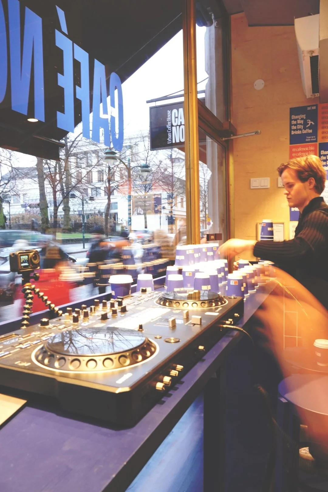 A DJ is mixing music at a cafe with two turntables and a digital controller in front of large windows. Outside, cars are passing by on a city street, and the cafe is illuminated with indoor lighting.