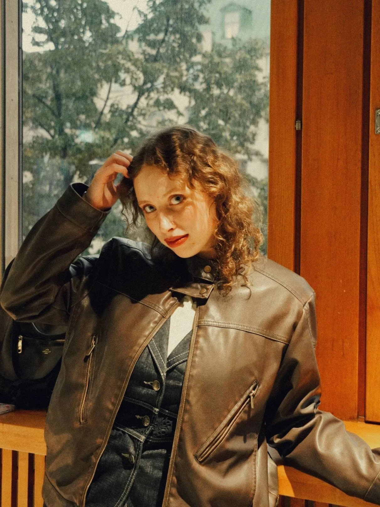 A woman with curly brown hair and red lipstick is sitting indoors near a wooden wall and a window with a tree outside. She is wearing a brown leather jacket and a dark outfit underneath. Stanislava Moskvina - Co-Founder of Symphony of views