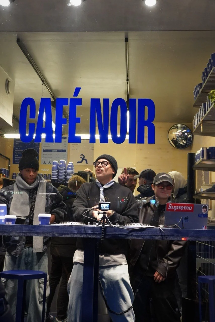 People standing behind the counter at Café Noir, with a large blue sign overhead, some of them in casual attire including beanies and sunglasses, with a mirrored wall and shelves of cups in the background.