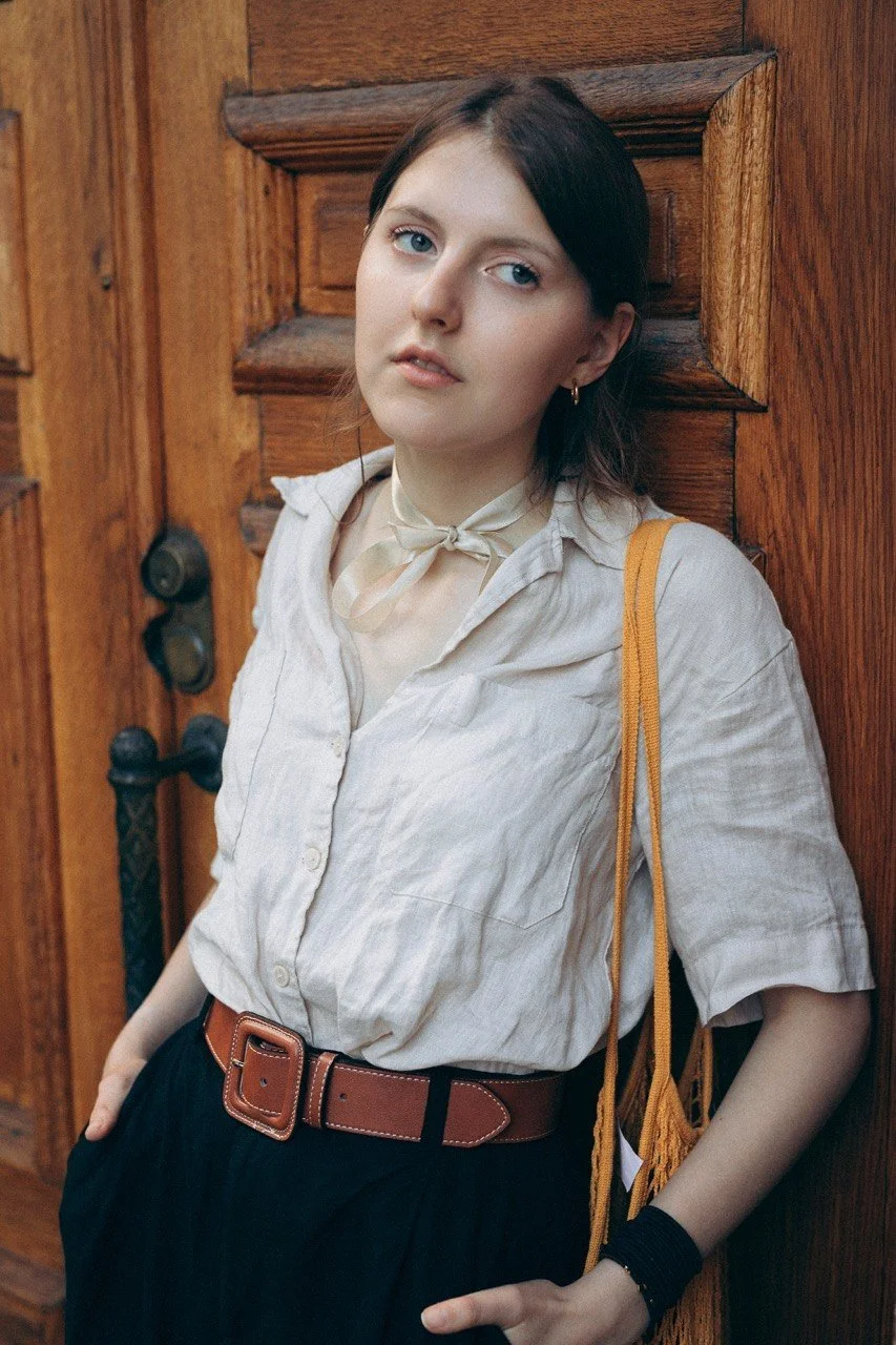 A young woman with fair skin and dark brown hair, wearing a cream-colored blouse with rolled sleeves, a beige neck scarf, and a brown belt, leaning against a wooden door with a serious expression. - Anna Krasnolobova - Founder of Symphony of Views