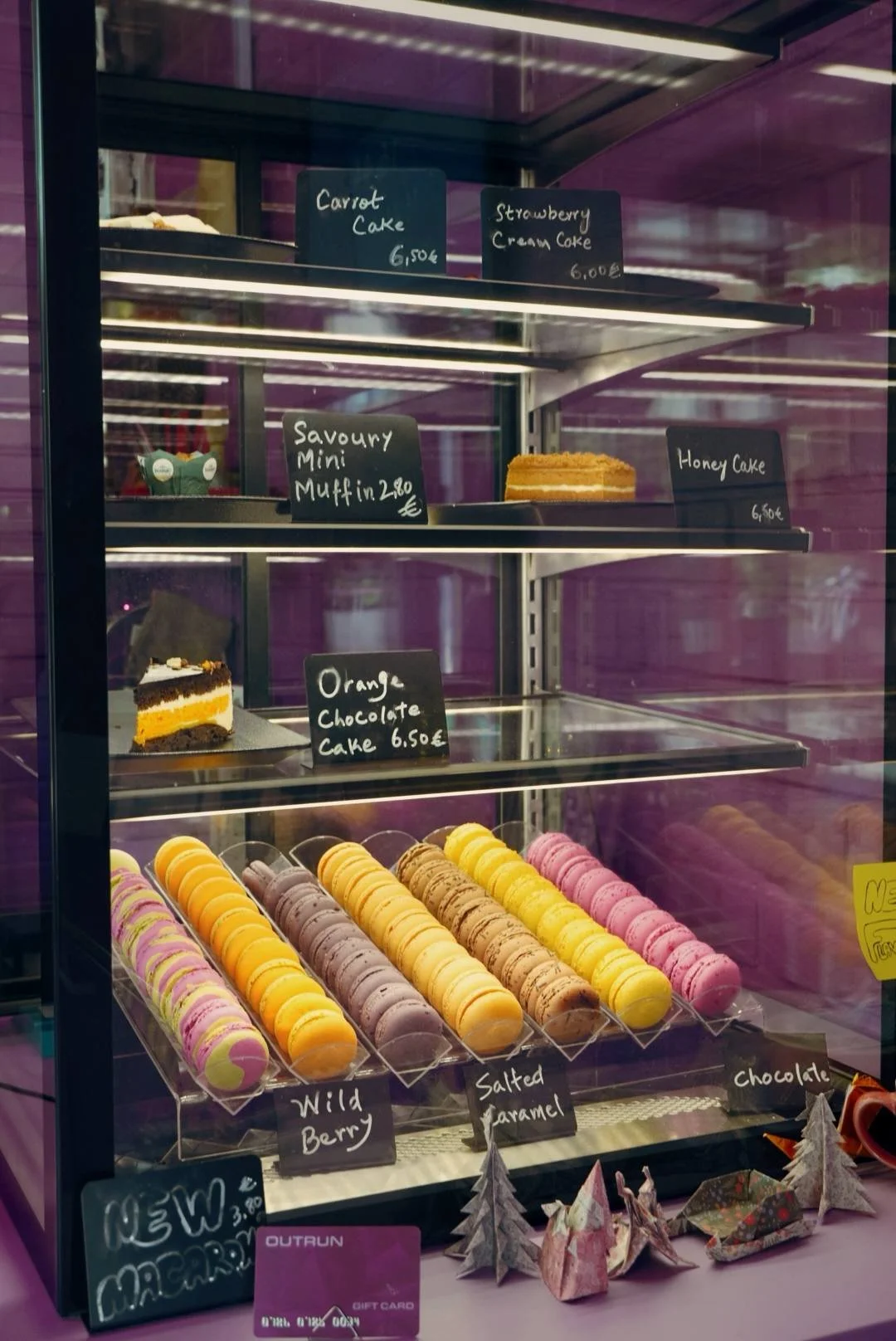 Display case with various cakes and macarons. Cakes include carrot, strawberry cream, honey, and orange chocolate. Macarons are in flavors like wild berry, salted caramel, and chocolate.