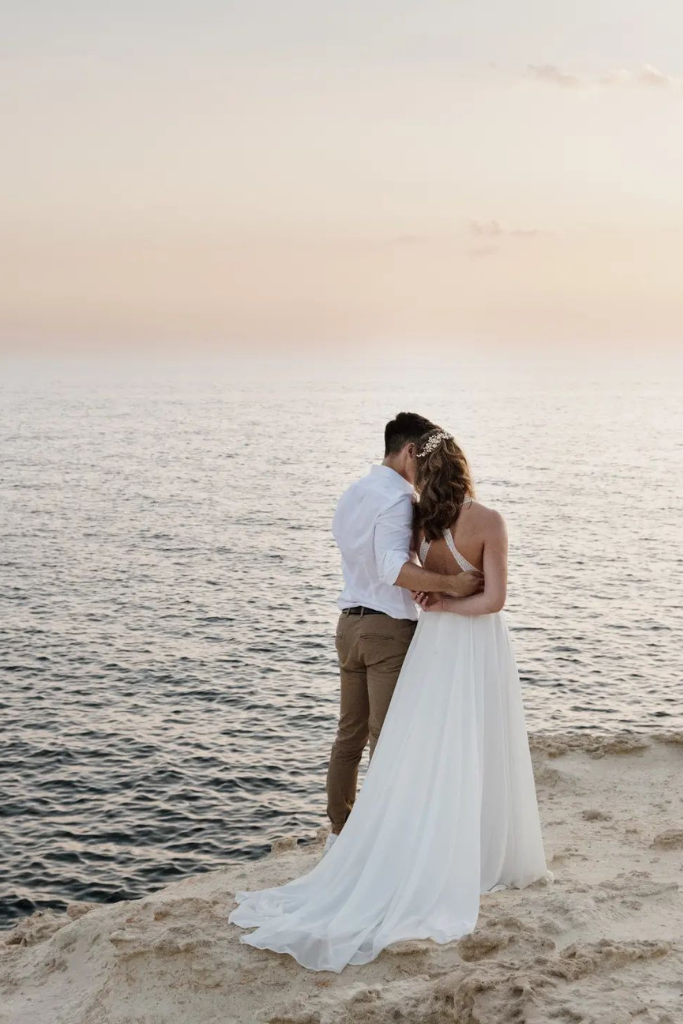 Un couple en tenue de mariage sur la plage, en train de s'embrasser au coucher du soleil.