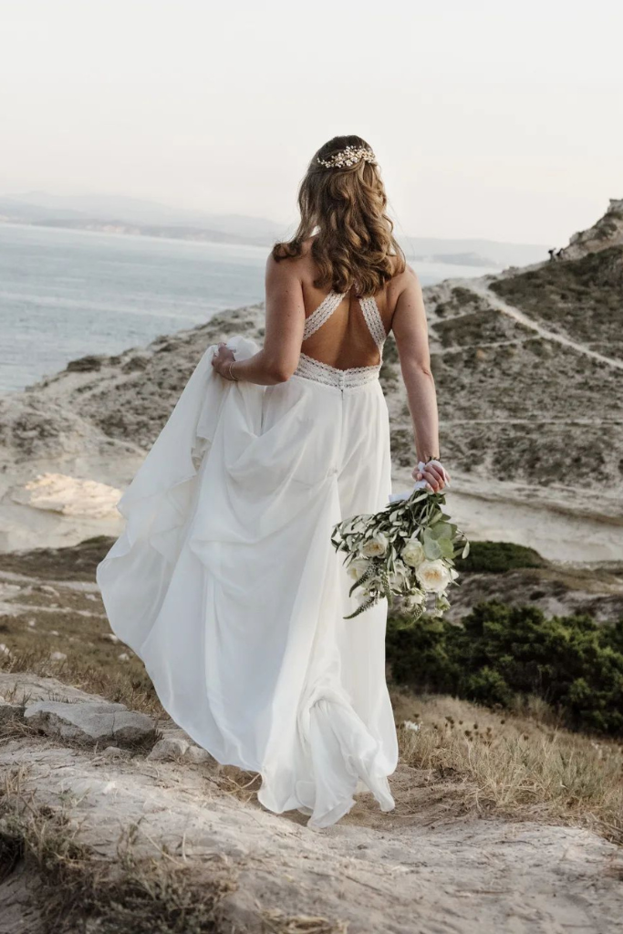 Femme en robe de mariée blanche avec couronne florale, tenant un bouquet de fleurs, marchant sur un chemin de terre près de la mer, avec des collines en arrière-plan.