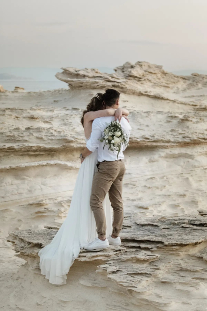 Un couple en tenue de mariage s'embrasse sur une falaise rocheuse au bord de la mer, avec paysage rocheux en toile de fond.