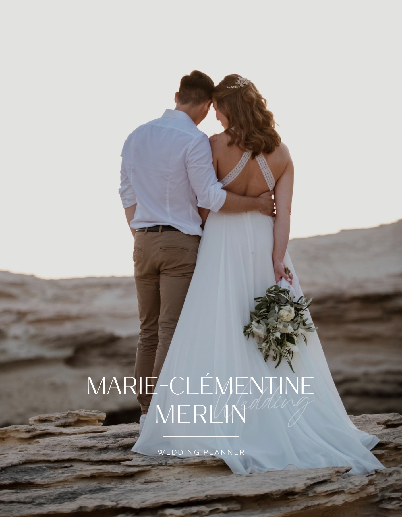 Un couple de mariés se tenant la tête en posant sur une plage lors du coucher du soleil, portant une robe de mariée blanche et un costume beige, avec un bouquet de fleurs à la main.