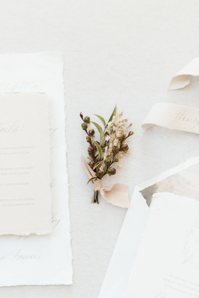 Arrangement floral avec petits bourgeons, feuilles vertes et ruban rose pâle, posé sur du papier blanc avec papiers à décor de mariage ou invitation.