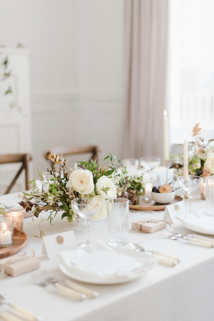 Table élégamment dressée avec une nappe blanche, vase de fleurs blanches et verts, bougies, assiettes, verres, couverts, et petits cadeaux dans des emballages beige.