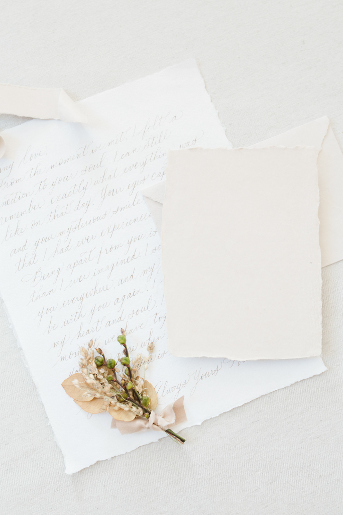 Une lettre manuscrite légèrement froissée, accompagnée d'une petite branche de fleurs et de feuilles séchées, attachée avec un ruban en satin beige. La lettre est posée sur un fond clair et neutre.