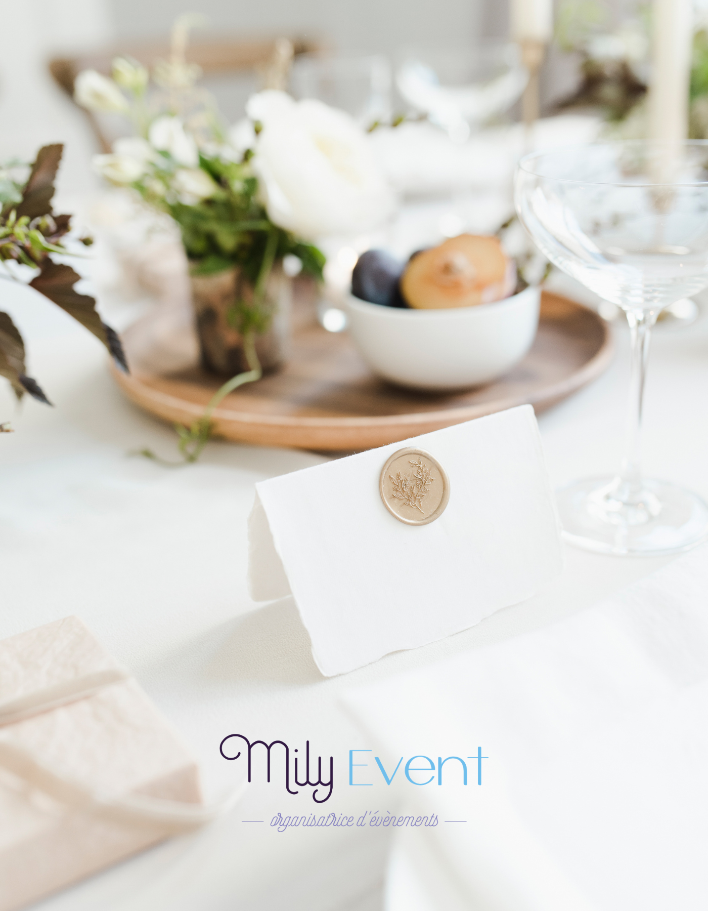 Table élégamment dressée avec des fleurs blanches, un bol de fruits, un verre à cocktail vide, et une enveloppe en cire sur une nappe blanche. L'image contient également un logo "Mily Event" avec la mention "organisatrice d'événements".